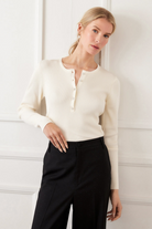 Woman wearing a cream sweater and black pants against a white wall