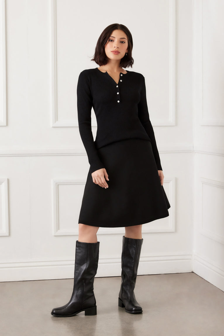 Woman wearing a black outfit with knee-high boots against a white paneled wall.