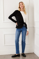 Woman wearing a black cardigan, white top, blue jeans, and black loafers against a white paneled wall.