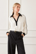 Woman wearing a white blouse with black accents and black pants against a white wall.