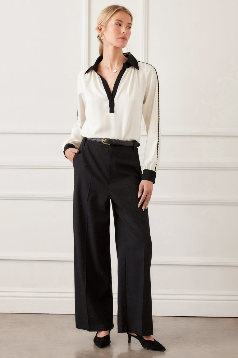 Woman wearing a white blouse with black trim and black pants against a white wall.