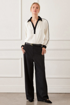 Woman wearing a white blouse with black trim and black pants against a white wall.