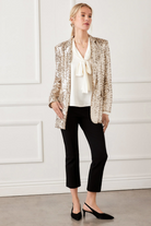 Woman wearing a gold sequin blazer over a white blouse with a bow, black pants, and black shoes against a white wall.