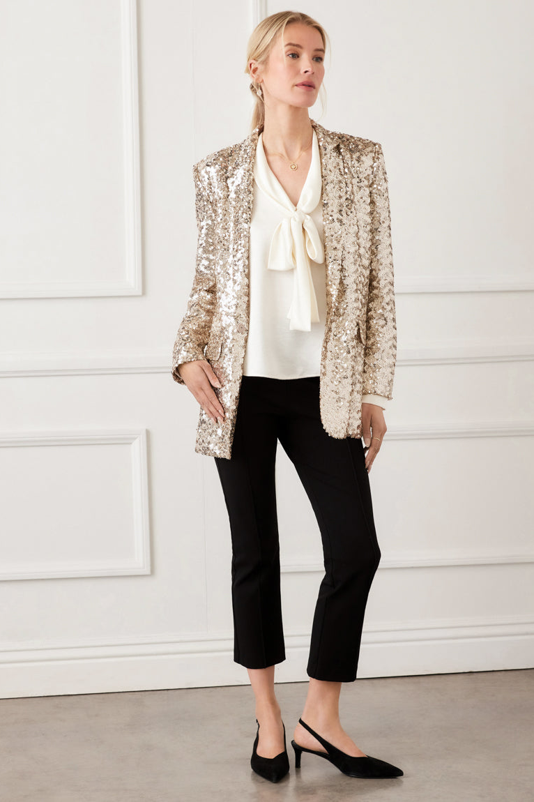 Woman wearing a gold sequin blazer over a white blouse with a bow, black pants, and black shoes against a white wall.