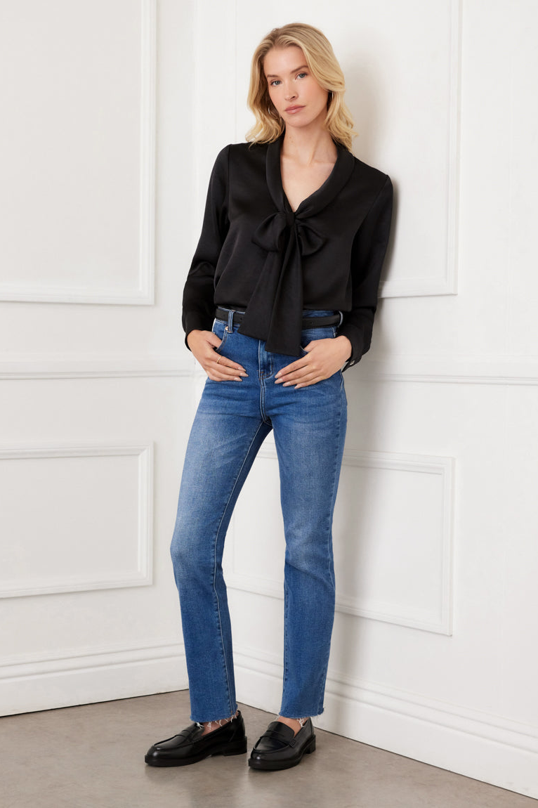 Woman wearing a black blouse with a bow tie and blue jeans against a white paneled wall.