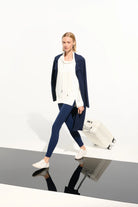 Woman in navy coat and blue leggings standing next to a white suitcase on a reflective surface.