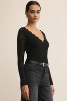 Woman wearing a black lace long-sleeve top and black pants against a beige background