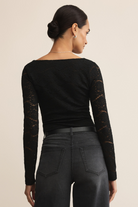 Woman wearing a black long-sleeve top with lace details and black pants on a beige background