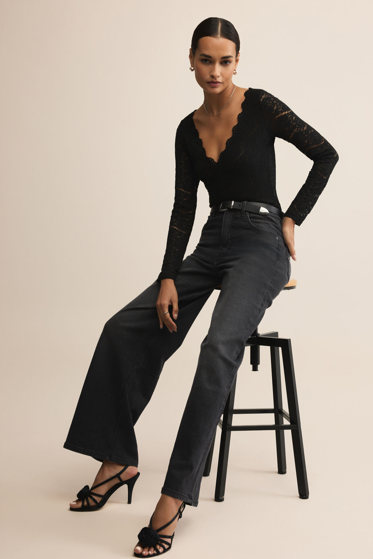 Woman wearing a black long-sleeve top and high-waisted dark jeans, standing on a stool against a beige background.