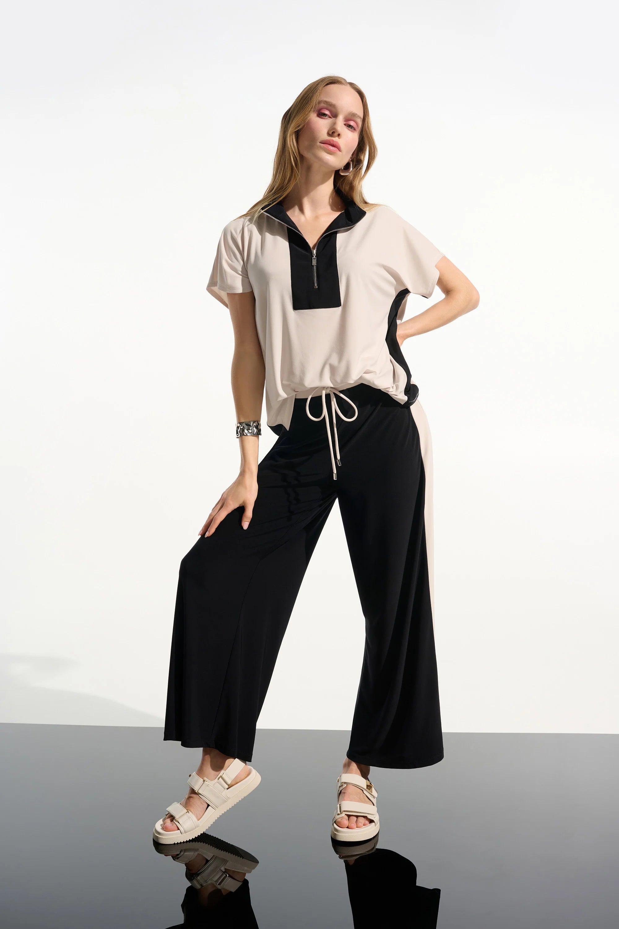 Woman wearing a white blouse with black accents and black pants on a reflective surface.