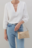 Person wearing a white sweater and blue jeans, holding a beige handbag against a plain background