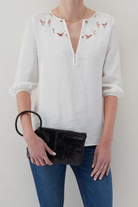 Person wearing a white blouse with floral embroidery and holding a black clutch against a plain background