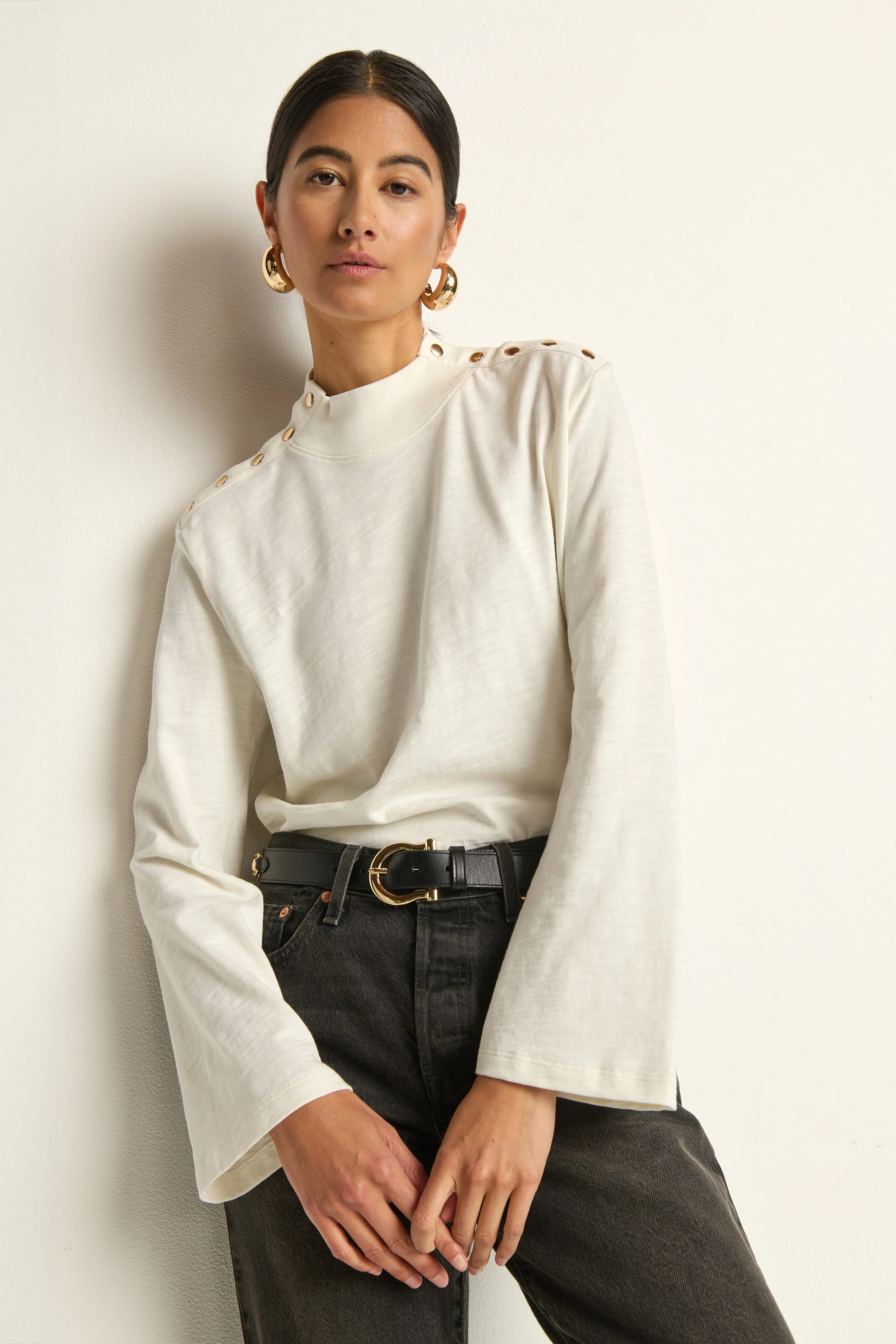 Woman wearing a white long-sleeve top with decorative buttons and black pants against a plain background