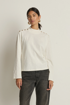 Woman wearing a white long-sleeve top with decorative elements and dark pants on a plain background