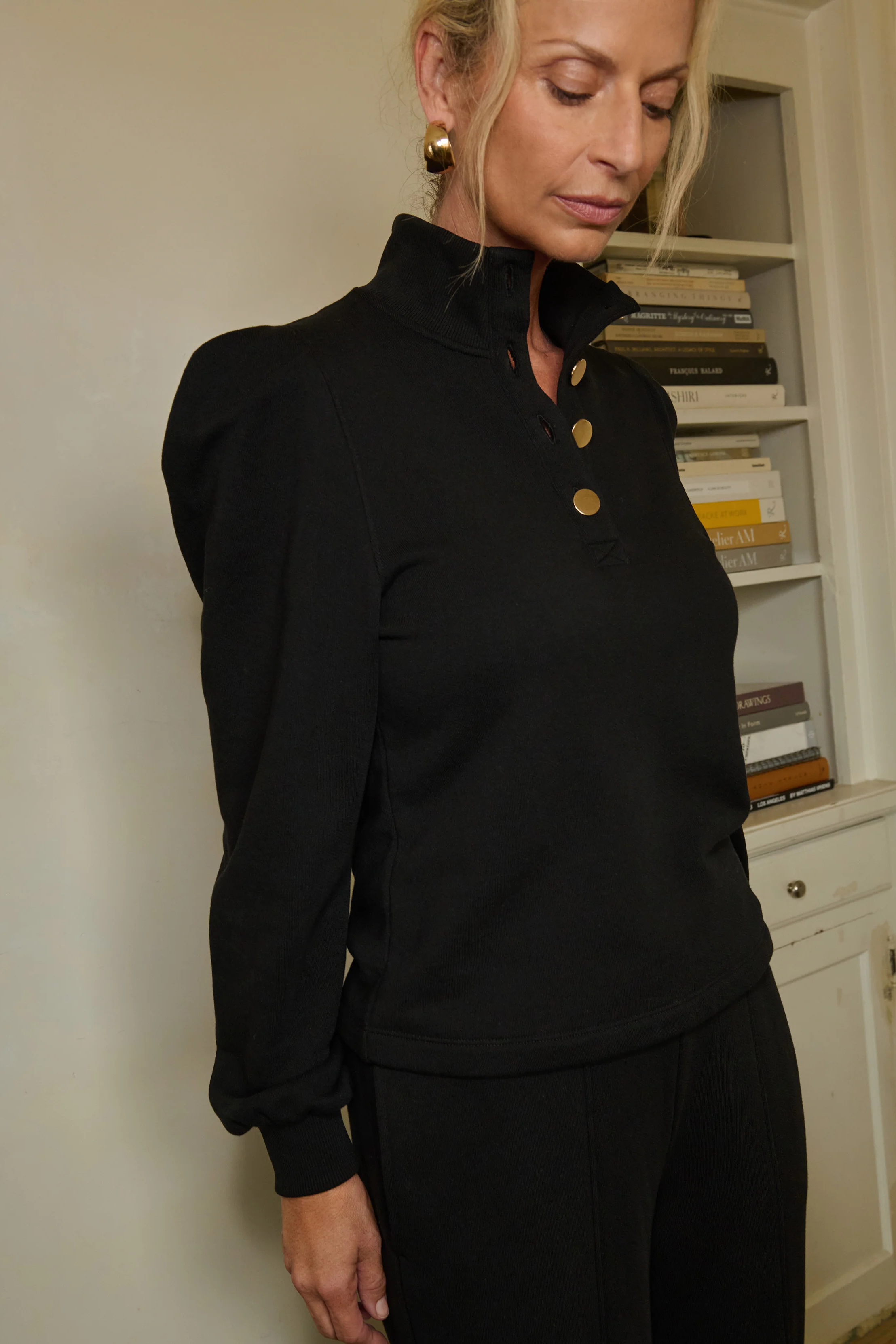 Woman wearing a black long-sleeve top with buttons in a room with a bookshelf.