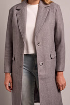 Person wearing a gray coat over a white shirt and dark jeans on a plain background