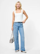 Woman wearing a white sleeveless top and light blue jeans, holding a gray handbag on a white background
