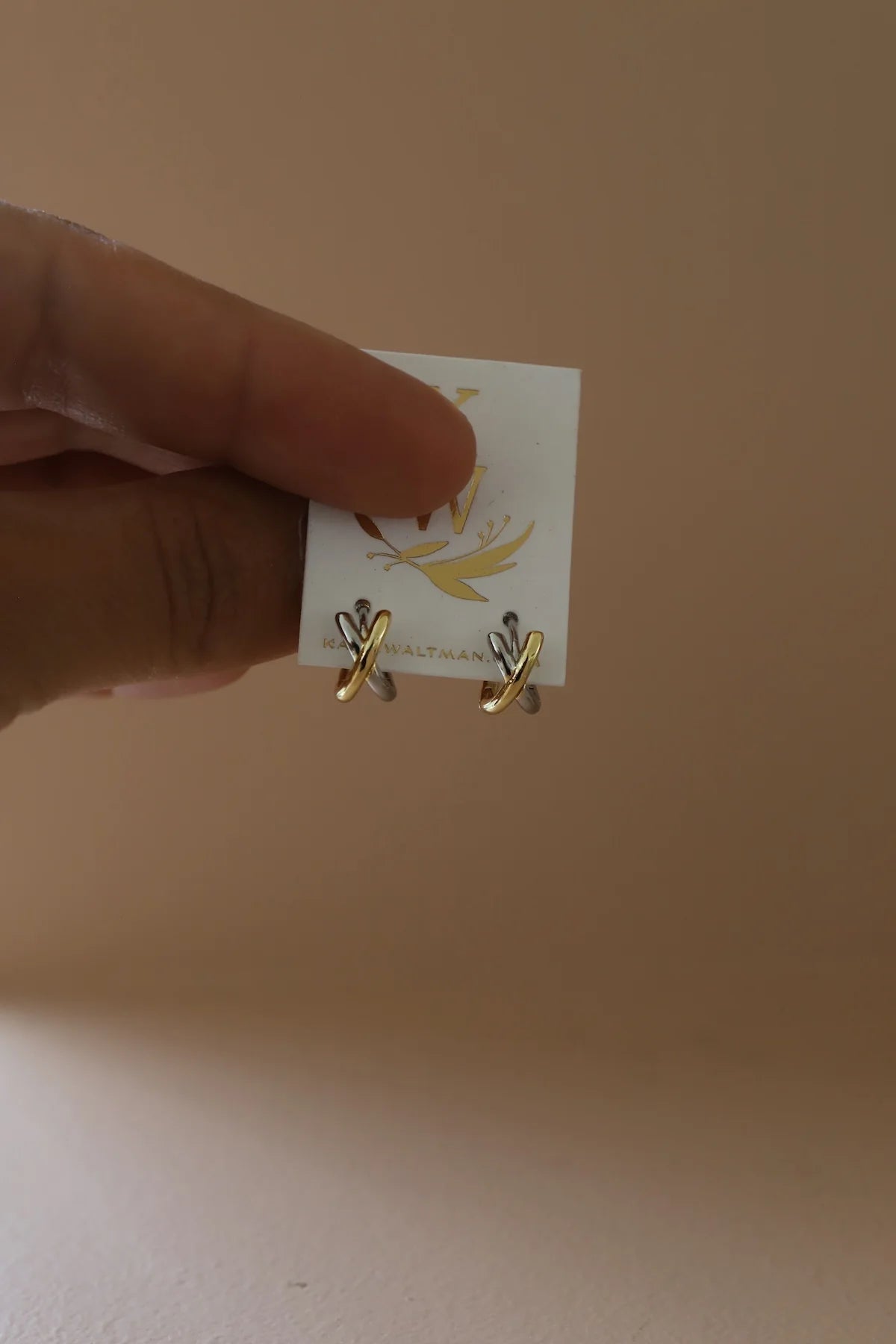Gold earrings held above a white card with a brand logo on a beige background