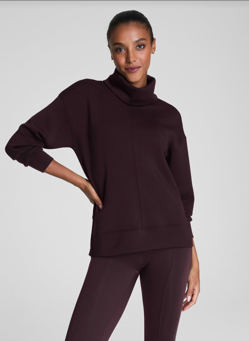 Woman wearing a dark brown turtleneck sweater and pants on a light gray background