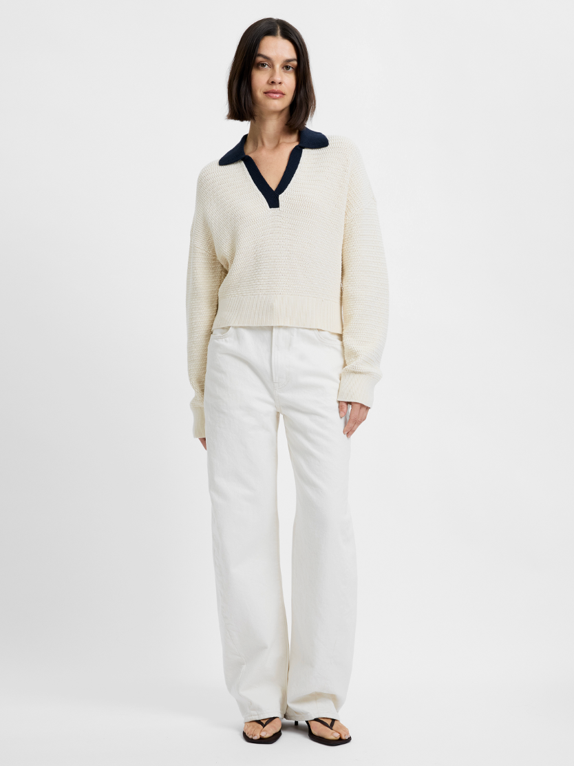 Woman wearing a cream sweater with a dark collar and white pants on a white background