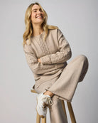 Woman wearing a beige cable knit sweater and pants sitting on a wooden stool against a plain background