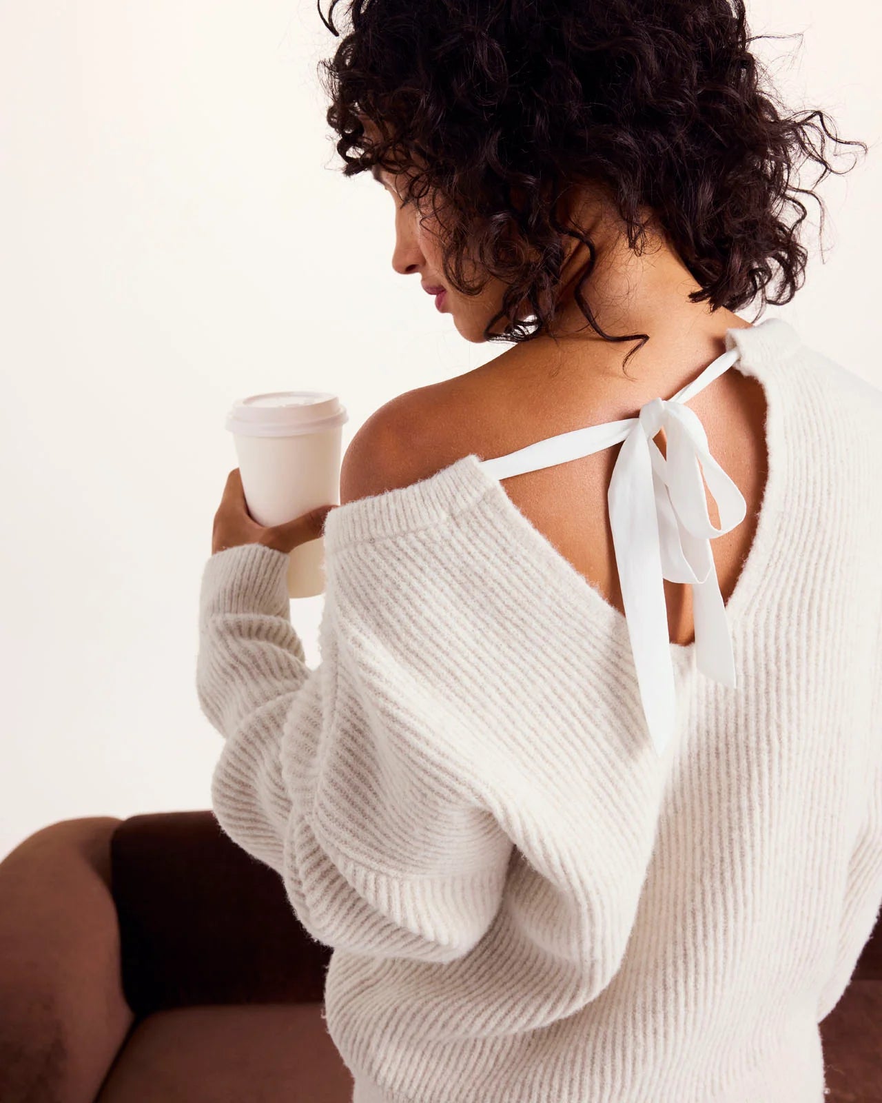 Woman wearing a white sweater with a bow detail, holding a coffee cup.