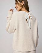 Woman wearing a beige sweater with a white bow detail on a light gray background