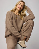 Woman wearing a brown sweater and pants sitting on a white stool against a light gray background