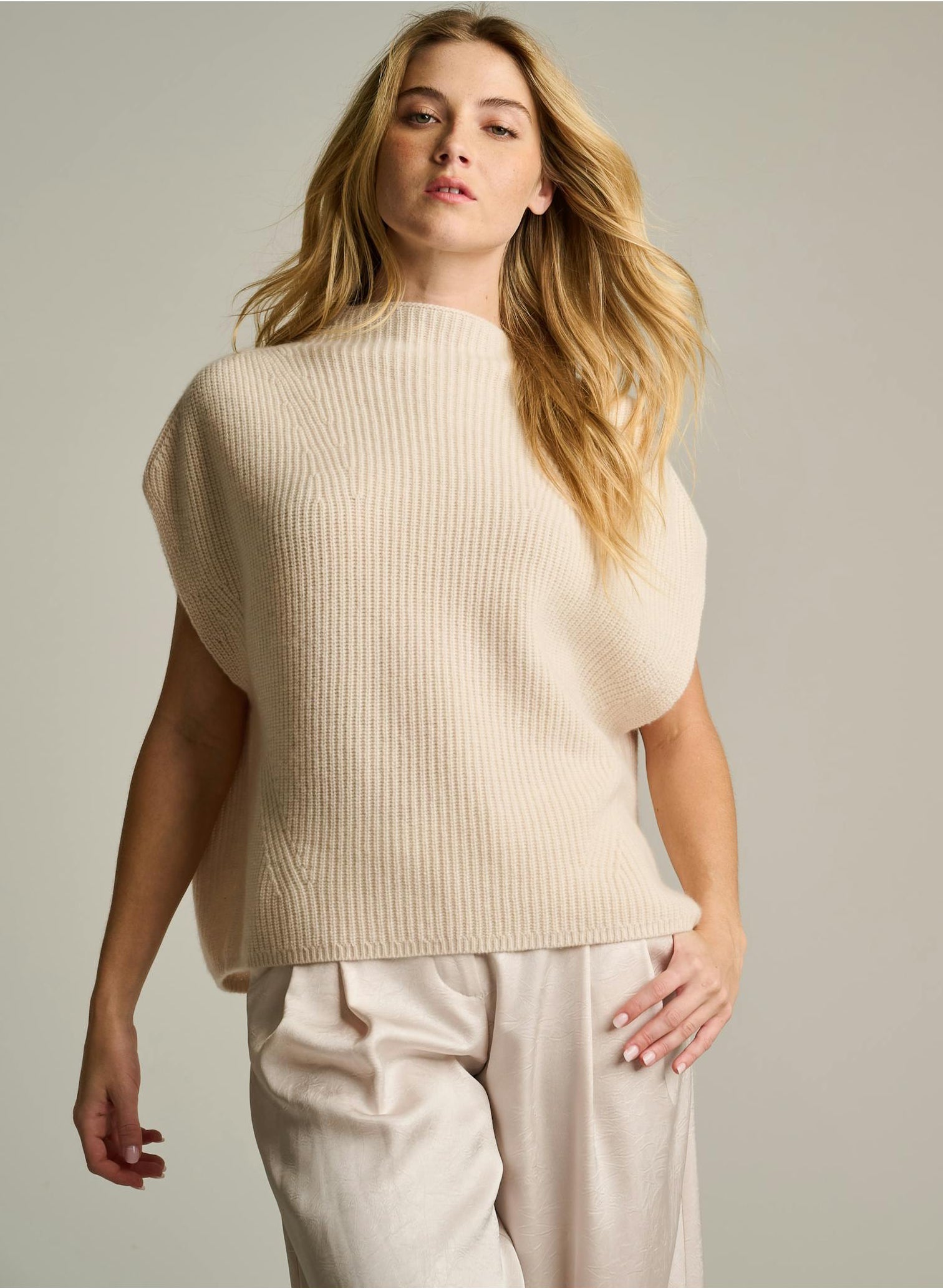 A woman modeling a sleeveless, ribbed knit sweater vest in a warm cream or off-white color, paired with matching light trousers, against a solid light gray studio background.